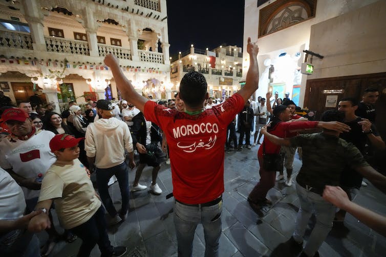 Un homme de dos, arborant un chandail où il est écrit ‘Morocco », salue une foule
