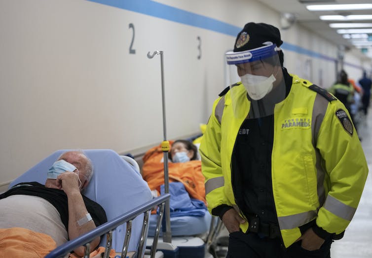 Un ambulancier portant une visière de protection et une veste jaune fluo longe une file de patients sur des civières dans un couloir d’hôpital