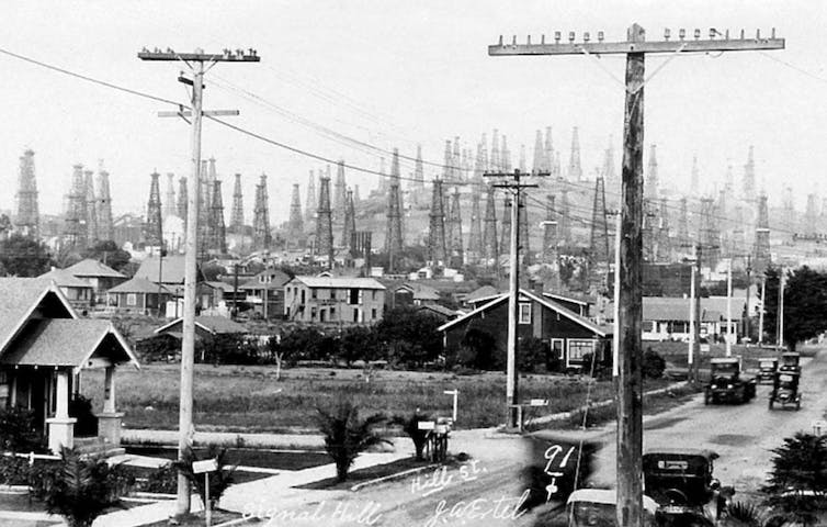 Une photo historique en noir et blanc montrant une rue avec des maisons, de vieilles voitures et des dizaines de derricks pétroliers sur la colline derrière elles
