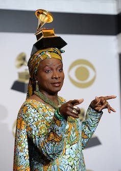Une femme en tenue africaine pointe ses doigts vers la pièce alors qu'elle pose pour une photo avec une statue en or d'un gramophone en équilibre sur sa tête.