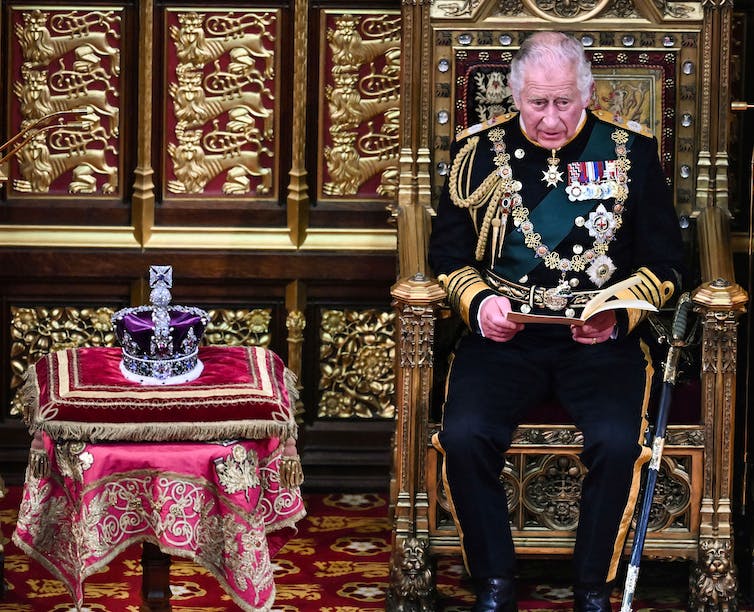 Un vieil homme sur un trône, assis à côté d’une couronne ornée de joyaux