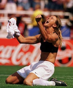 Une femme en soutien-gorge de sport et en shorts lève son chandail, les poigts serrés, en criant