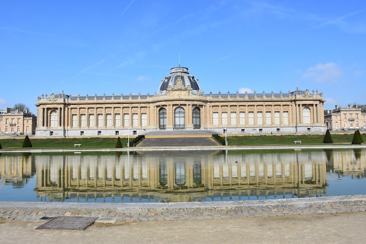 Un long plan d'un bâtiment beige avec son reflet dans un bassin d'eau.