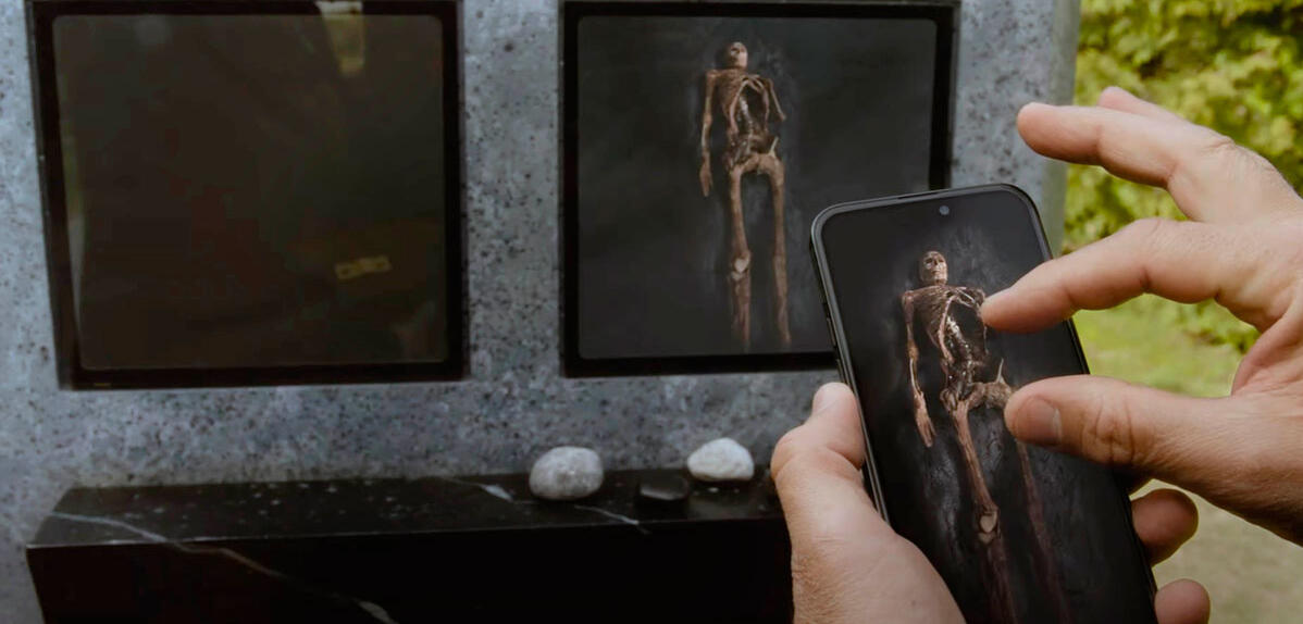 image du film où une main touche un téléphone qui montre l’intérieur de la tombe et on voit le squelette