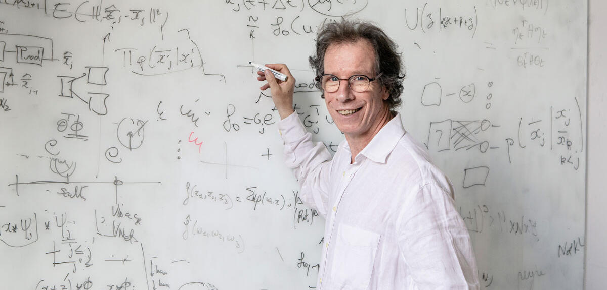 Portrait de Stéphane Mallat devant un tableau blanc couvert de formules mathématiques