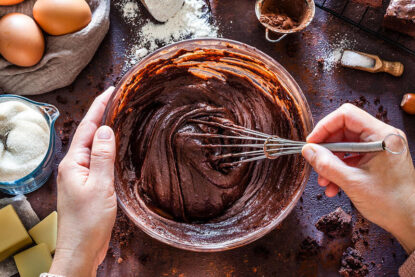 Des mains mélangent une pate chocolatée avec un batteur, les ingrédients du gâteau sont disposés autour