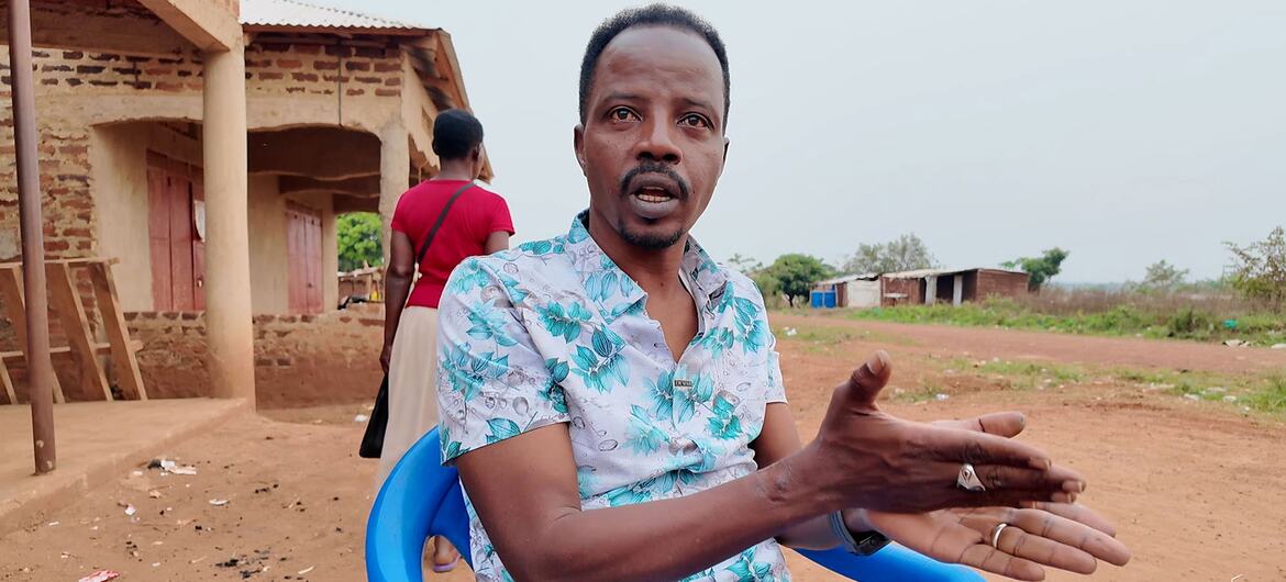Un homme à la moustache et aux cheveux courts, vêtu d'une chemise à fleurs, s'assoit sur une chaise bleue et fait des gestes en parlant. Derrière lui, une femme en chemise rouge passe devant un bâtiment en brique dans un cadre rural.