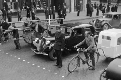 Dans une rue de Paris en 1940, un agent de police retient derrière une ligne de clous divers engins roulants
